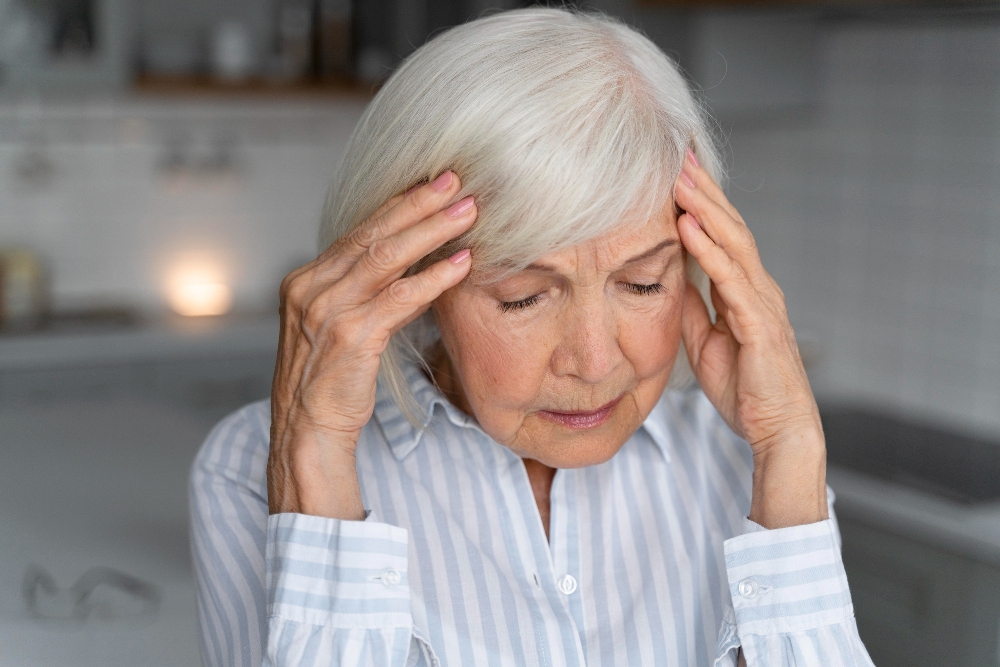 Elderly woman presses fingers to temples, eyes closed in kitchen.